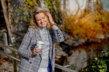 Uzun saçlı, açık gri kaşmir kazaklı ve uyumlu tüvit ceketli zarif sarışın güneşli bir sonbahar gününde parkta yürüyüş yaparken sıcak kahvenin tadını çıkarıyor. Sonbahar havası. Pozitif duygular. 