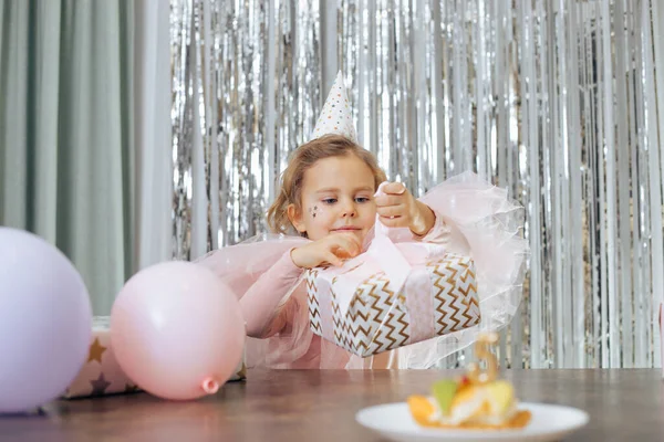 Doğum gününü kutluyoruz. Parti şapkalı ve pembe havalı kıyafetli çekici kızıl saçlı kız güzel tasarlanmış hediyeleri açma konusunda tutkulu. Balon ve pastanın yanında. Hediye teslimatı. Mutlu çocukluk. 