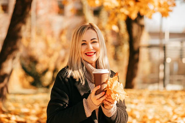 Gorgeous blonde lady in black cashmere coat with bouquet of orange leaves and cup of coffee is enjoying sunny autumn day in city park . Share joy. Walking in fresh air. Weekend. Beautiful weather.