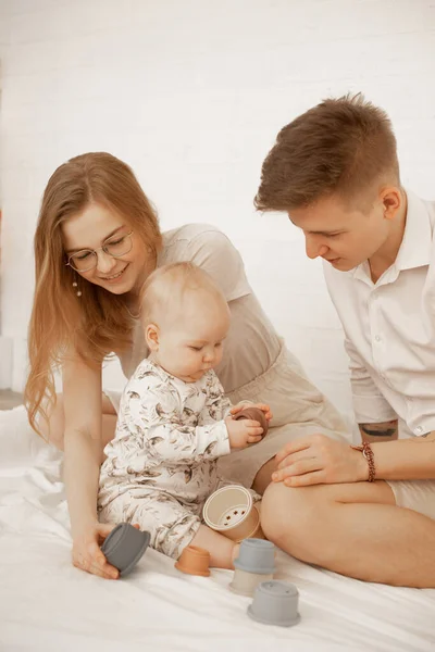 Portrait of young happy beautiful family with little cute plump infant baby playing with modern pastel silicone bowls of different size, building pyramid, sitting on white bed. Early development.