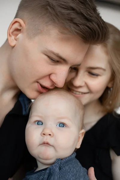 Portrait of young family in dark clothes with cherubic little plump grey-eyed baby infant toddler kissing on white background. Childhood, parenthood, motherhood, fatherhood, love, care, protection.