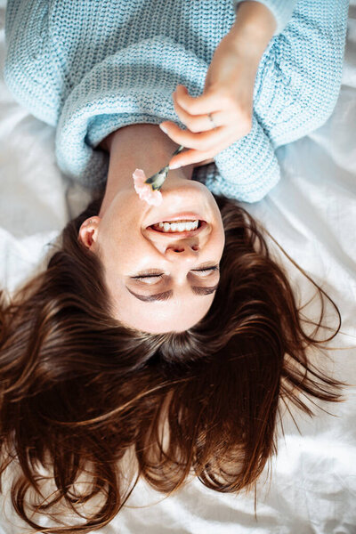 Милая молодая женщина в синем свитере с длинными темными волосами, лежащими на кровати с цветами и веселящимися. Природная красота, красивое обращение, образ жизни