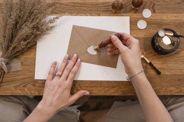 Beyaz balmumu mühür yapan, kahverengi bir zarf yapıştıran, ahşap masa başında kuru otlar, siyah balmumu ısıtıcısı ve kaşıklarla oturan, tanınmamış bir kadının iyi korunmuş ellerinin üst görüntüsü..