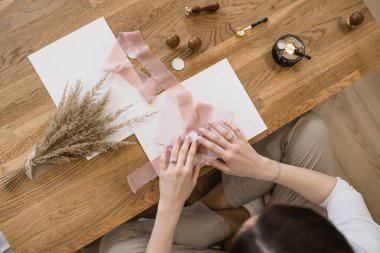 Beyaz karşılama zarfını pembe şeffaf kurdeleyle dekore eden, ahşap masa başında bir buket pampa otu, pullar, balmumu mühür, kaşık ve siyah balmumu ısıtıcısı ile oturan tanınmamış bir kadının üst görüntüsü..