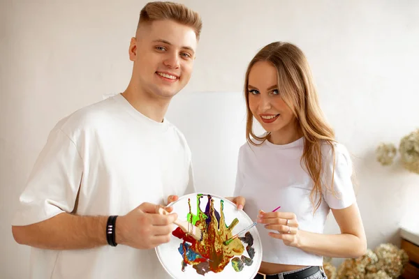 Married couple drawing together. Girlfriend and boyfriend spending time together, doing arts. Woman and man looking at camera and holding brush and pallette with colors. Two artists painting