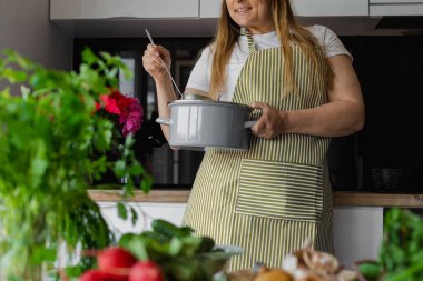Taze sebze salatası ve yeşillik kullanarak mutfakta yemek pişiren iri yarı, gülümseyen sarışın kadın. Vejetaryen yemeği ve yaşam tarzı. Turp, patates, salatalık, pancar ön planda..