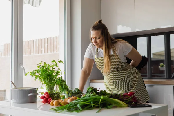 Plump, plus size blonde woman cooking healthy food in the kitchen, choosing fresh vegetable salad, greenery. Vegetarian food and lifestyle. Radish, potato, beetroot and sprouting onion on the table