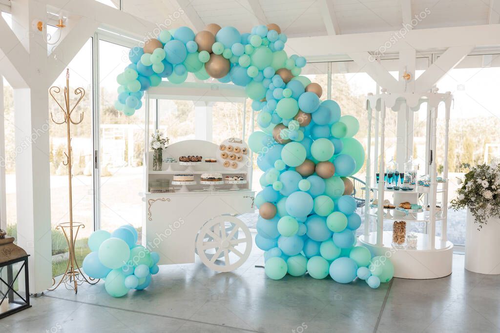 Festive stand with delicious desserts and fresh drinks, blue balloons arch in white banquet hall. Artwork, decorated hall, event celebration. Holiday, tasty food and decorations, beautiful service.
