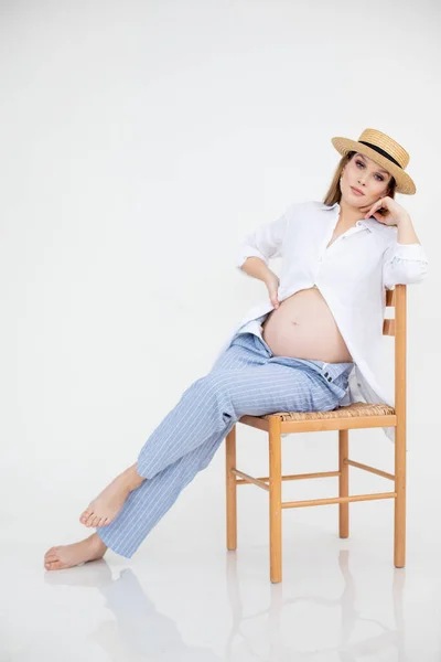 Pregnant woman in linen clothes and hat on white background sitting on chair. Pregnancy, maternity concept. Beautiful woman, pregnancy. Studio shot