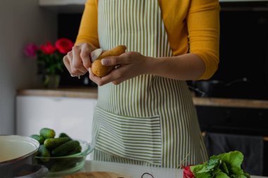 Önlüklü, patates soyan ve mutfaktaki tahta tahtada taze sebze doğrayan bir kadın. Karışık yaz salatası ya da çorba pişirmek. Salatalık, tavaya yakın pancar, masada öğle yemeği. Sağlıklı beslenme.