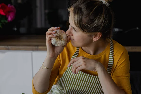 Closeup blonde concentrated woman in striped apron smelling fresh garlic bulb in kitchen. Cooking vegetable mixed salad, organic detox soup with condiment. Natural farm food. Diet and ecological dish