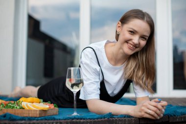Mavi ekoseli terasta beyaz şarap ve aperatiflerle uzanan mutlu genç kadın. Alkol içmek ve dışarıda eğlenmek. Ev partisi, tatil ve dinlenme odası..