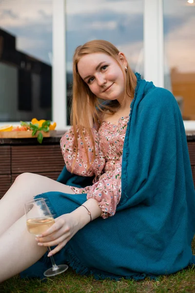 Happy young woman wrapped in blue plaid sitting on grass terrace with glass of white wine and snacks closeup. Drinking alcohol drink and enjoying outdoor recreation. Home party, vacation and lounge.