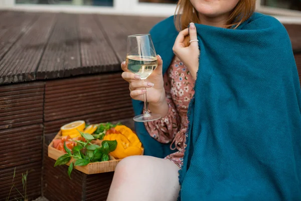 Woman wrapped in blue plaid sitting on terrace with glass of white wine and plate of snacks closeup. Drinking alcohol drink and enjoying outdoor recreation. Home party, vacation and lounge.