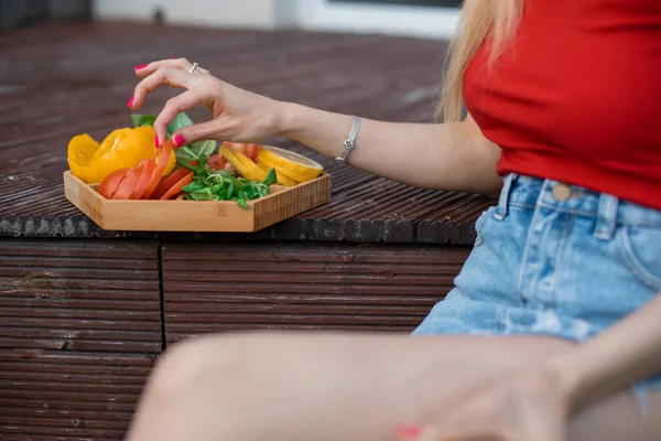 Woman sitting on terrace and eating fresh greens, fruit and vegetables from wooden plate closeup. Snack composition and healthy food. Enjoying outdoor recreation. Home party, vacation and lounge.