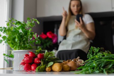 Masada taze sebzeler var. Artı önlüklü sarışın kadın mutfakta akıllı telefon kullanıyor, arka görüntüde indeksleme yapıyor. Mobil uygulama tarifi, yeni yaz yemeği fikri. Diyet ve sağlıklı organik yemek.