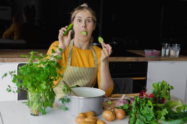 Ağzında salatalıkla kameraya bakan komik ve şaşırmış sarışın kadın salata için taze sebze pişiriyor. Patates, yeşillik, tavanın yanında pancar. Sağlıklı beslenme, diyet ve yaz tarifi