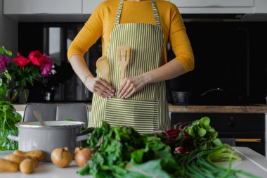 Çizgili mutfak önlüğü içinde tanınmayan bir kadın, cebine ahşap spatula çatalı koyuyor. Kadın aşçı yemek yapmaya başlar, yemek tarifini düşünür. Organik karışık salata için taze sulu vitamin sebzeleri.
