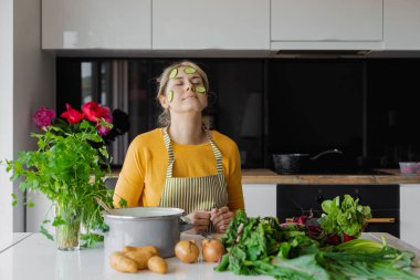 Mutfakta maske niyetine yüzüne salatalık dilimlenmiş komik, gevşemiş sarışın kadın. Cilt bakımı için taze sebzeler. Salata ve doğal organik ekolojik meyve suyu. Evde tedavi ve kozmetoloji