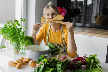 Yüze yakın ahşap mutfak spatulası ekipmanlarını geçen komik sarışın kadın. Taze organik yemek pişirmek için çatal bıçak kullanmak. Pan ve yaz sulu sebzeler masada. Salata için malzeme seçin