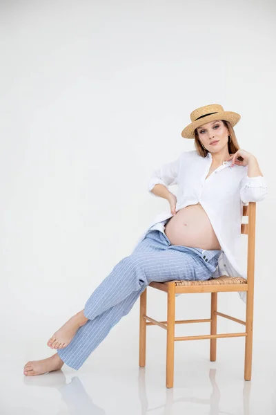 Pregnant woman in linen clothes holds hands on belly sitting on chair on white background. Natural beauty, stylish pregnancy, studio shot