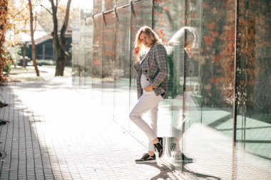 Ceketli ve açık renk pantolonlu güzel sarışın cam vitrine yaslanıp düşünüyor. Camdaki kadın yansıması. Moda ve güzellik. Sonbahar manzarası.