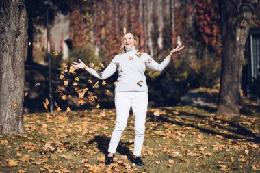 Yaprak düştü. Açık renk elbiseli gülümseyen sarışın güneşli bir günde şehir parkına yaprakları atarken eğleniyor. Olumlu duygular. Doğayla birlik. Sonbahar manzarası. Mutlu anlar. 