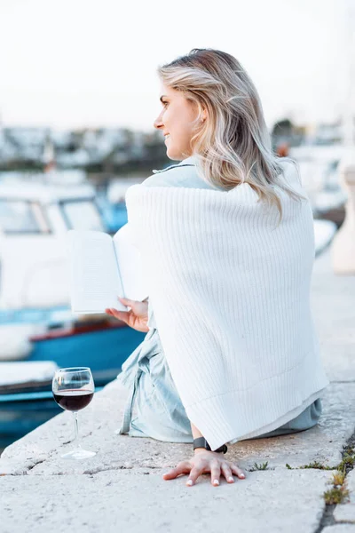 Young woman with wavy loose hair is sitting on pier against background of sea and yachts with book in her hands, there is glass of red wine next to her. Tourism and recreation.