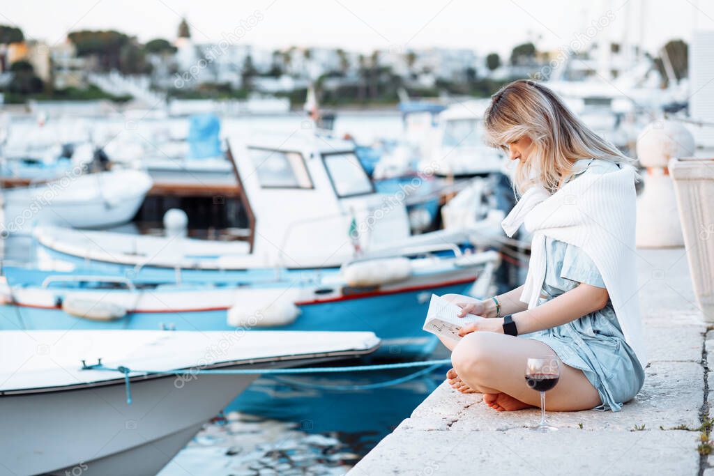 Young woman is sitting cross-legged on pier and reading book against background of sea and yachts, glass of red wine is standing next to her. Boat trip on yacht. Happy moments.