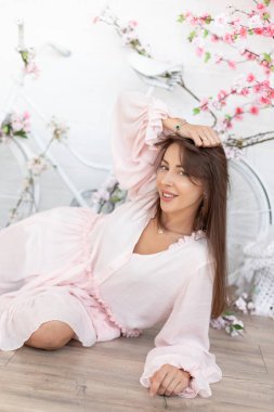 Romantik soluk pembe elbiseli, kahverengi saçlı çekici genç kadın, çiçek tomurcuklarıyla süslenmiş beyaz bisikletin arka planında, ahşap zeminde yatıyor. Romantik ruh hali.
