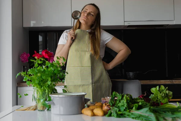 Plump, plus size blonde woman smelling tasty soup in pan with close eyes, holding ladle near face. Summer vitamin recipe for diet, use potato, beetroot, greenery. Cook healthy organic eco food at home