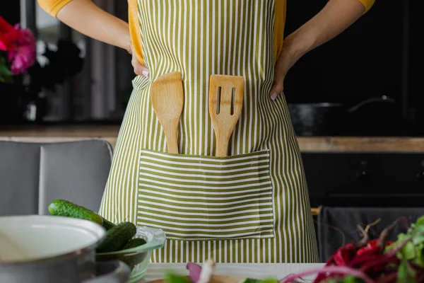 Unrecognizable woman with arms akimbo in striped kitchen apron with wooden spatula utensil inside. Chef or cook at home. Thinking what recipe to choose. Fresh vitamin vegetables, ingredients on table