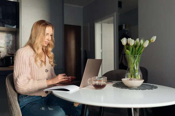 Attractive young woman with long wavy hair in large-knit pink jacket and jeans works online at home in her kitchen at round white table. Accepting orders online. Online training. Meeting online.