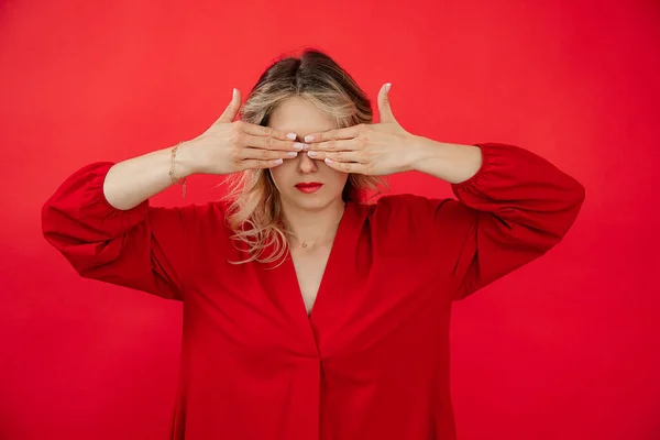 Portrait of hiding woman in red outfit with perfect bright makeup closing eyes by hands isolated on red background. Avoiding problems, fear, giving under stress. Fashion and beauty industry.