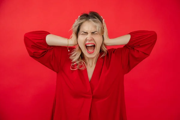 Portrait of loudly screaming woman in red outfit with perfect bright makeup holding on head by hands isolated on red background. Losing control, giving under stress. Fashion and beauty industry.