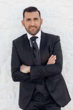Siyah takım elbiseli, kısa siyah saçlı yakışıklı nişanlısının portresi, çapraz elli beyaz duvarda duruyor. Düğün, tören, özel etkinlik, kutlama. Dikey.