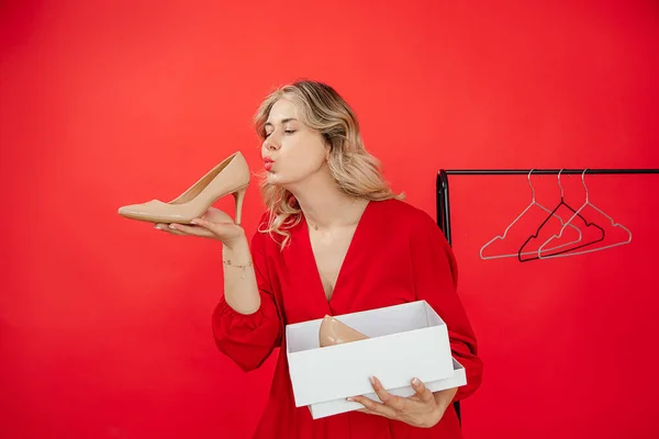 Awe young caucasian woman holding patent leather high heels beige shoes on red background with copy space, happy, kissing. Sales, shopping, stylist, wardrobe, clothes hanger