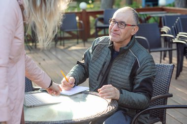 Siyah ceketli, gözlüklü orta yaşlı bir adamın portresi, açık bir kafede masada oturuyor, elinde turuncu kalemle not defterine notlar yazmaya hazır, kadına bakıyor. Eğitim, iletişim.