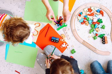 Küçük çocuklar ve eğitimciler lego katlayıp renkli kalemlerle çiziyorlar oyun odası manzarasında yerde oturuyorlar. Yaratıcılık ve zekayı geliştiren anaokulu öğrencileri için ilginç bir ders..