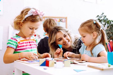 Küçük kızlar ve gülümseyen eğitimci geometrik figürleri çalışıyor, oyun masasının üzerinde oyun oynuyor ve katlıyor oyun odasında yerde oturuyor. Mantık ve zeka geliştiren anaokulu öğrencileri için ilginç bir ders..