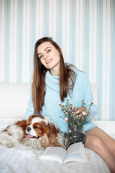 Blue-eyed young brown-haired woman in loose pale blue sweater is smiling happily sitting with her beloved dog Kavler King Charles spaniel on bed with book. Positive emotions. Owners and pets.