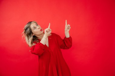 Parlak kırmızı elbiseli güzel bir kadının portresi başını öne eğiyor ve kırmızı arka planda izole edilmiş parmakları işaret ediyor. Modern reklam, bedava fotokopi alanı. Moda ve güzellik endüstrisi.
