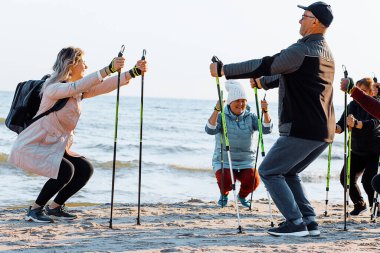 Koç olarak gülümseyen kadın, deniz kıyısında profesyonel sopalarla veya direklerle İskandinav yürüyüşü öğretiyor. Yaşlıları eğlendirmek, nefes almak için yürüyüş eğitimi alıyor. Seyahat etkinliği
