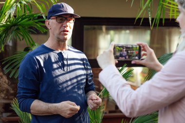 Kafedeki palmiye ağaçlarında duran kendinden emin orta yaşlı blog yazarı portresi, blog için içerik oluşturuyor, deneyimi paylaşıyor, kadının elinde tuttuğu akıllı telefonla eğitim videosu dersleri kaydediyor..