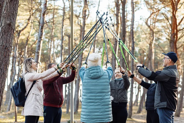 Group of people different ages making Nordic walking with trekking sticks in the forest among trees. Crossed sticks as symbol of teamwork. Prepare to sport trip education of senior and mature people