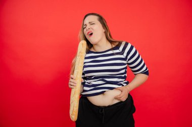 Çizgili tişörtlü, üzgün, şişman bir kadının mikrofon olarak Fransız ekmekli baget tutması ve kırmızı stüdyo arka planında çıplak göbeğine dokunması. Tatillerde kilo alırsın. Boşluğu kopyala