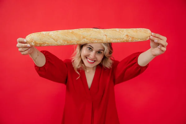 Smiling glad and joyful blonde woman in red dress holding and showing long french bread baguette in hands on red studio isolated background. Promotion of snacks, tasty, delicious food for the market