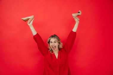 Memnun ve gururlu kırmızı gömlekli, kolları yukarıda kırmızı arka planda kırmızı ayakkabılı sarışın bir kadın. Yaz alışverişinin ve büyük satışların tadını çıkar. Kıyafet ve aksesuar al.