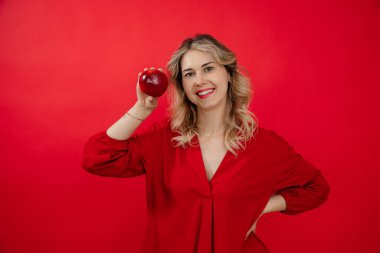 Genç, neşeli, neşeli, mutlu, sarışın kadın yüzünün yanında taze elma tutuyor, kırmızı izole edilmiş arka planda güzel beyaz bir gülümseme sergiliyor. Sağlıklı yaşam tarzı, sağlıklı beslenme. Beyazlatma ve diş temizleme