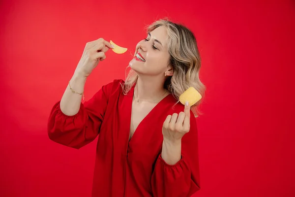 Smiling glad and joyful blonde woman in red dress holding and eating potato chips, crisps in hands on red studio isolated background. Promotion of food, snacks, tasty, delicious food for the market